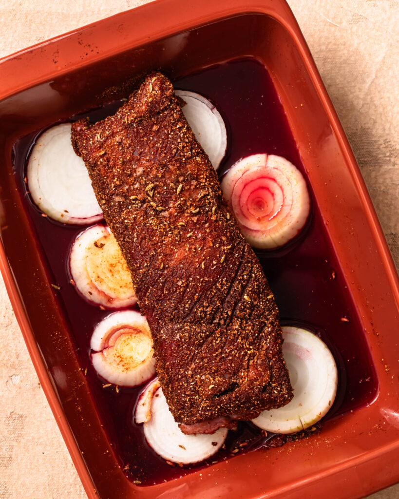 Beef brisket coated in spice rub sitting atop sliced onions and pomegranate juice. 