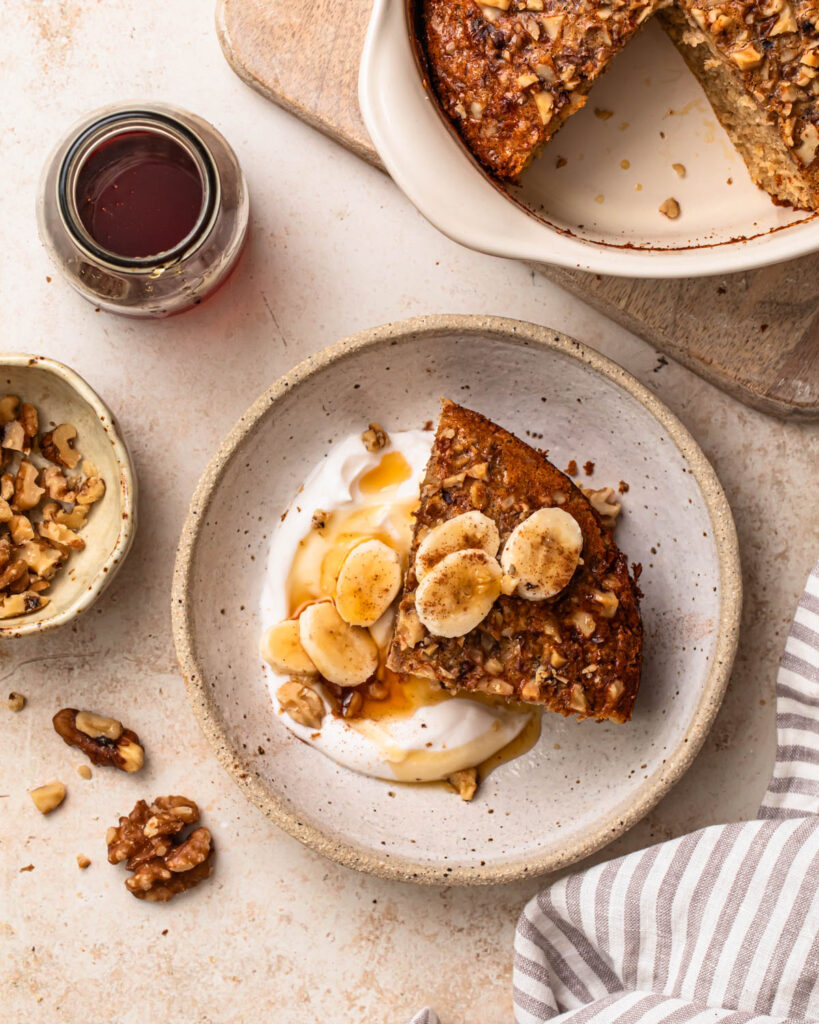 A slice of banana bread baked oats in a bowl with yoghurt, banana slices and maple syrup.