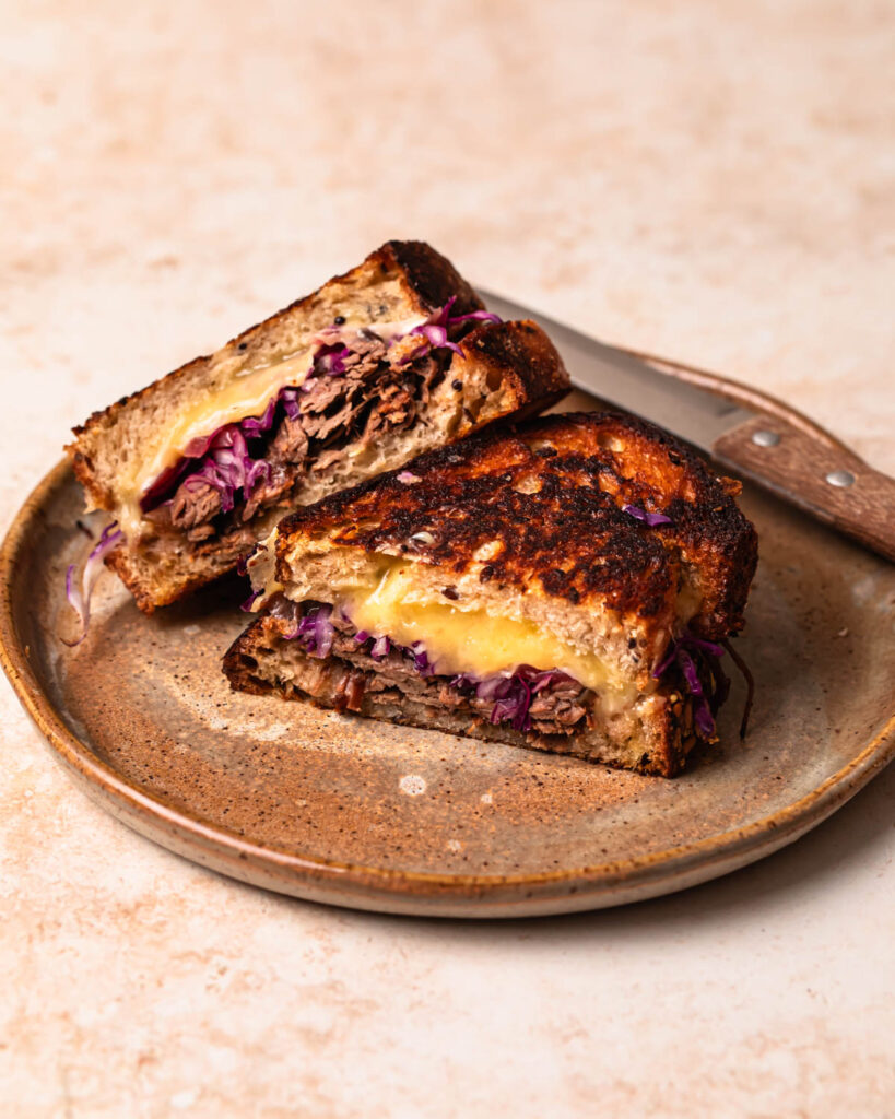 A brisket grilled cheese sliced in half, with one half leaning on the other on a plate.