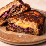 A brisket grilled cheese sliced in half, with one half leaning on the other on a plate.