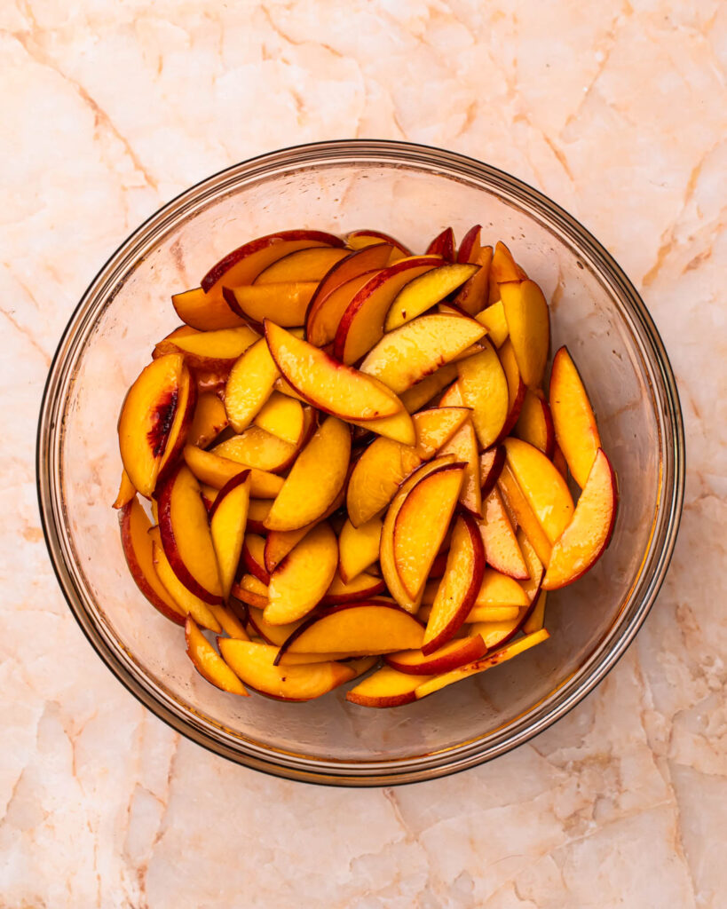 Thinly sliced peaches in a glass mixing bowl with maple syrup and rose water.