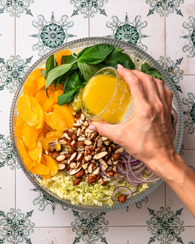 Salad dressing being poured over a citrus slaw in a glass bowl. 