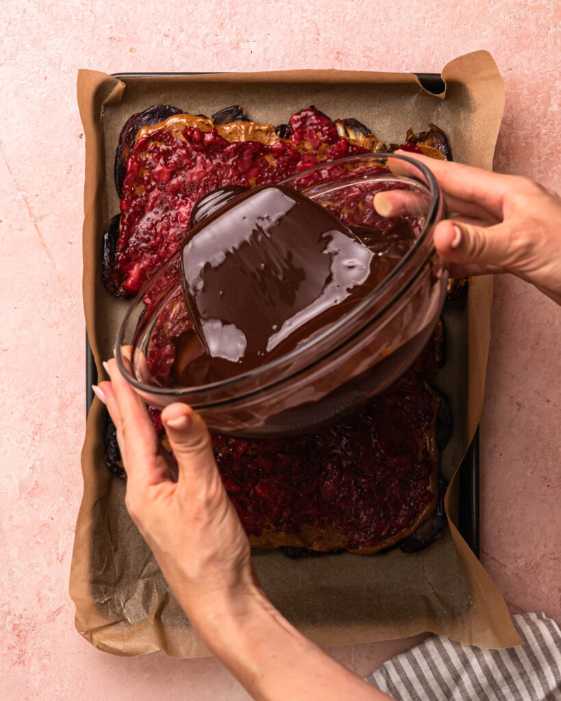 Dark chocolate being poured onto the layer of medjool dates, almond butter and strawberry chia jam. 