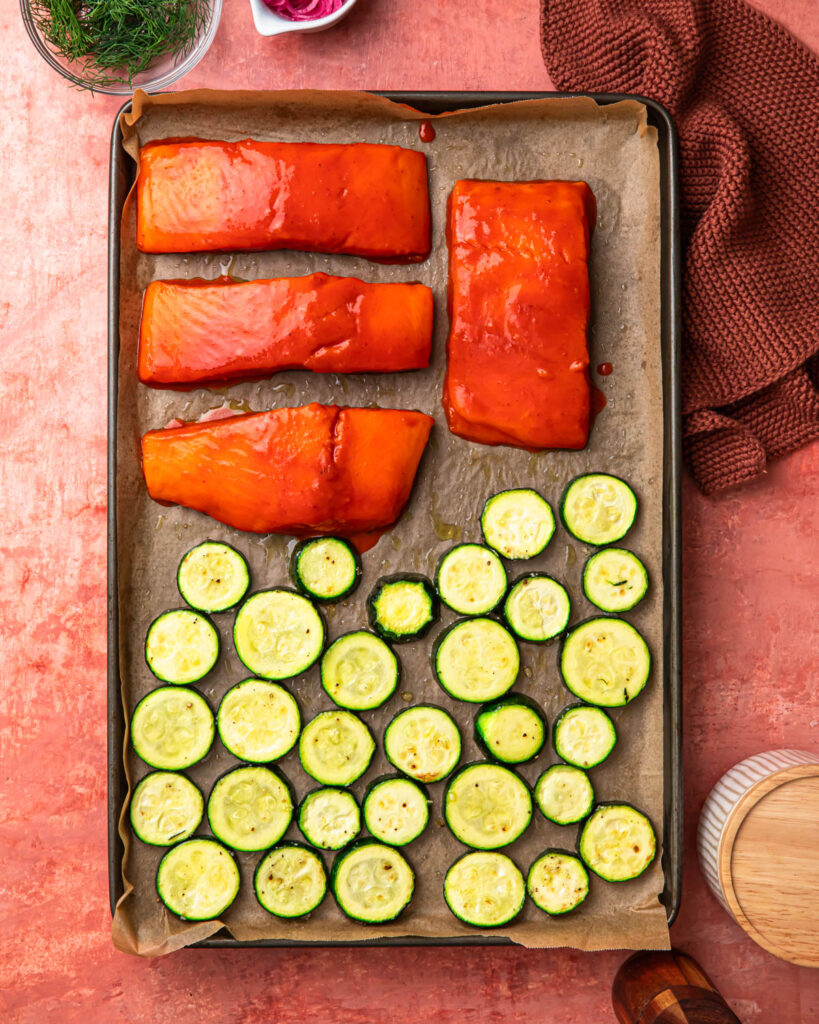 Harissa honey marinaded salmon added to a tray with partially cooked zucchini.