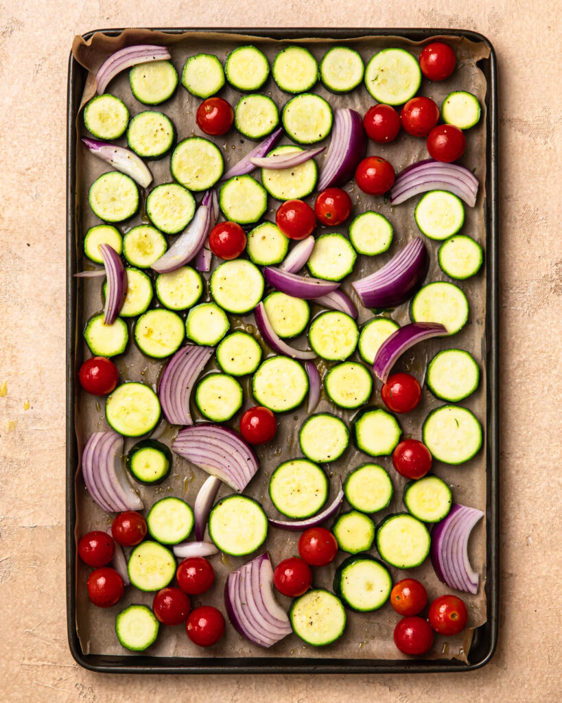 Seasoned zucchini rounds, cherry tomatoes and onion wedges spread out into a single layer on a baking tray.