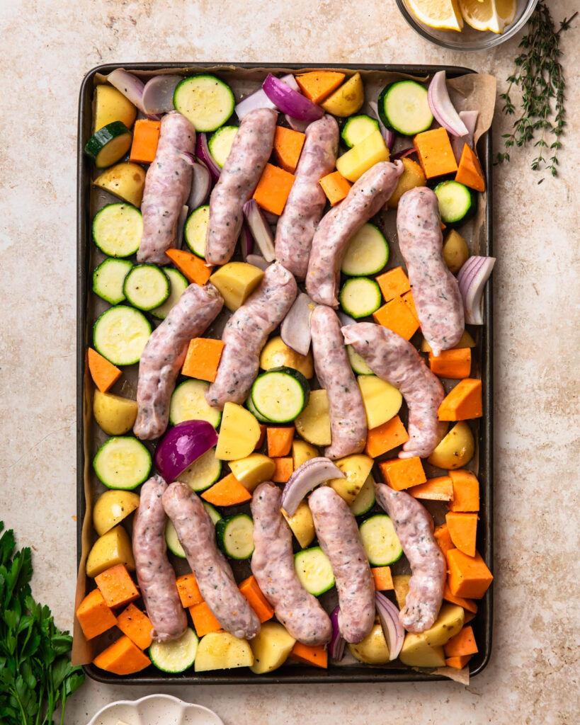 Seasoned vegetables spread out on a sheet pan with chicken sausages nestled into the vegetables. 