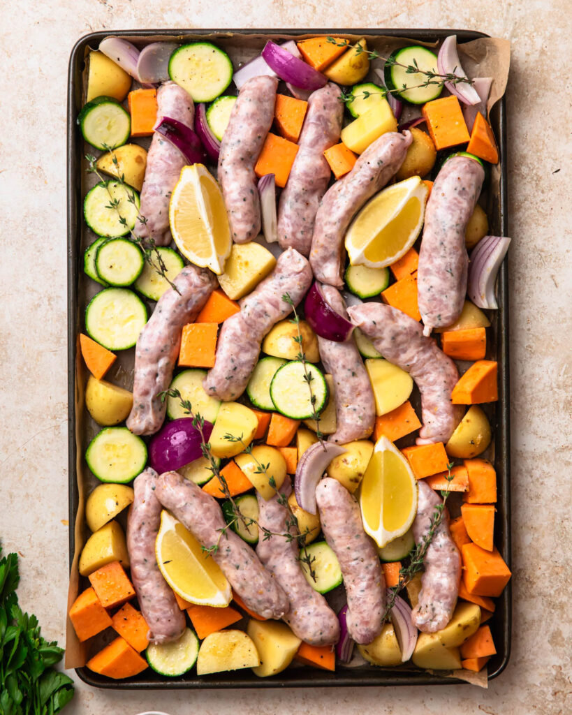 Uncooked sheet pan chicken sausage and vegetables spread out on a sheet pan and topped with lemon wedges and thyme.