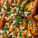 Close up photo of sheet pan chicken sausages and vegetables cooked on a pan and topped with parsley and feta.