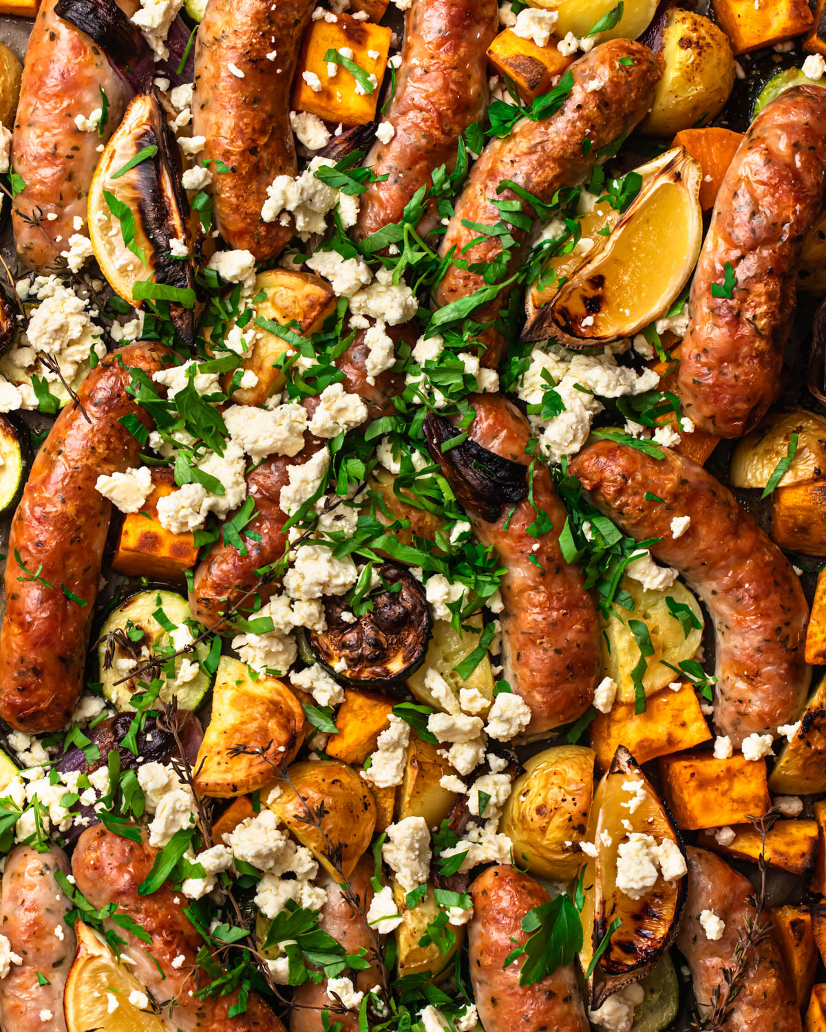 Close up photo of sheet pan chicken sausages and vegetables cooked on a pan and topped with parsley and feta.