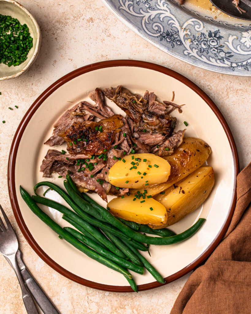 Slow cooked lamb shoulder served on a plate with lemony potatoes and steamed green beans.