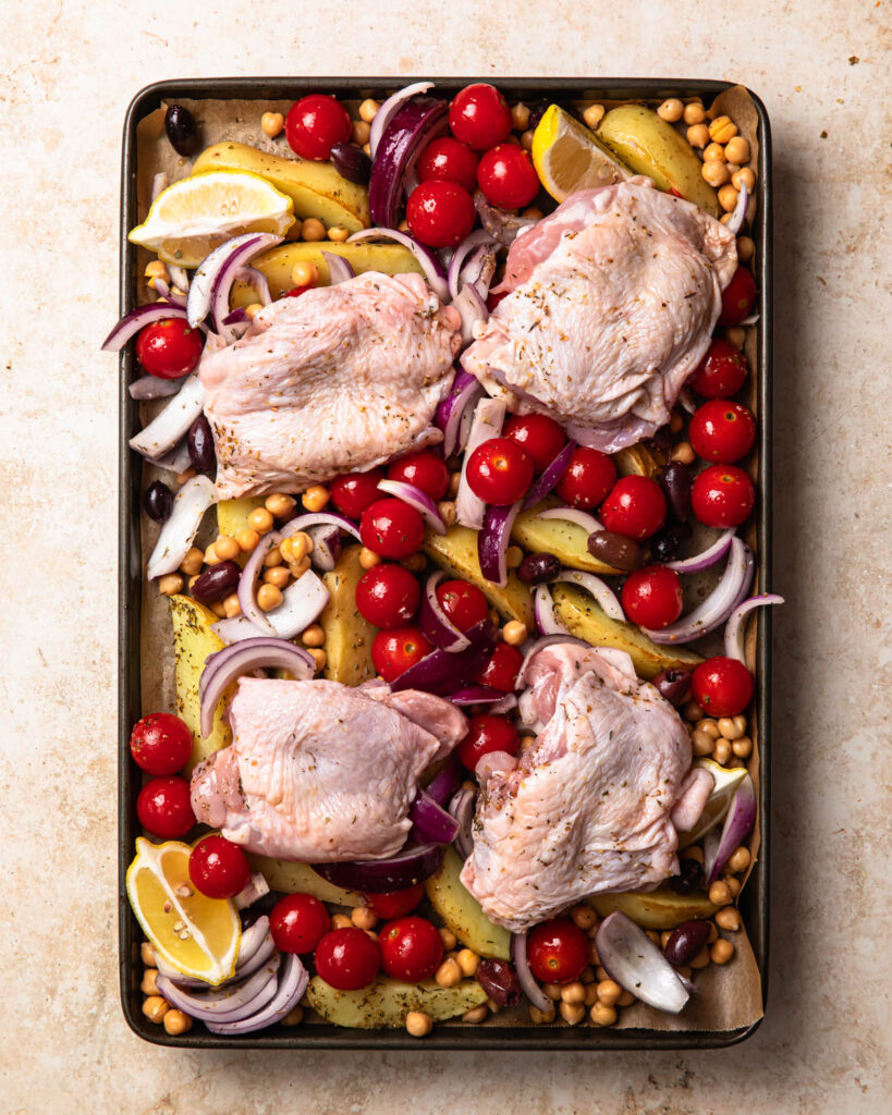 Marinaded chicken thighs added to a large sheet pan with other vegetables to make Mediterranean sheet pan chicken.