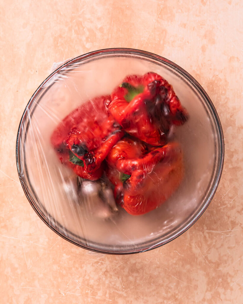 Roasted red peppers and garlic cloves in a glass bowl covered with plastic wrap.