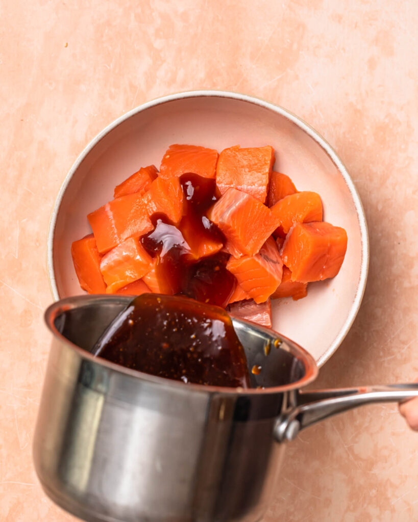 Teriyaki sauce being poured onto salmon cubes in a bowl.