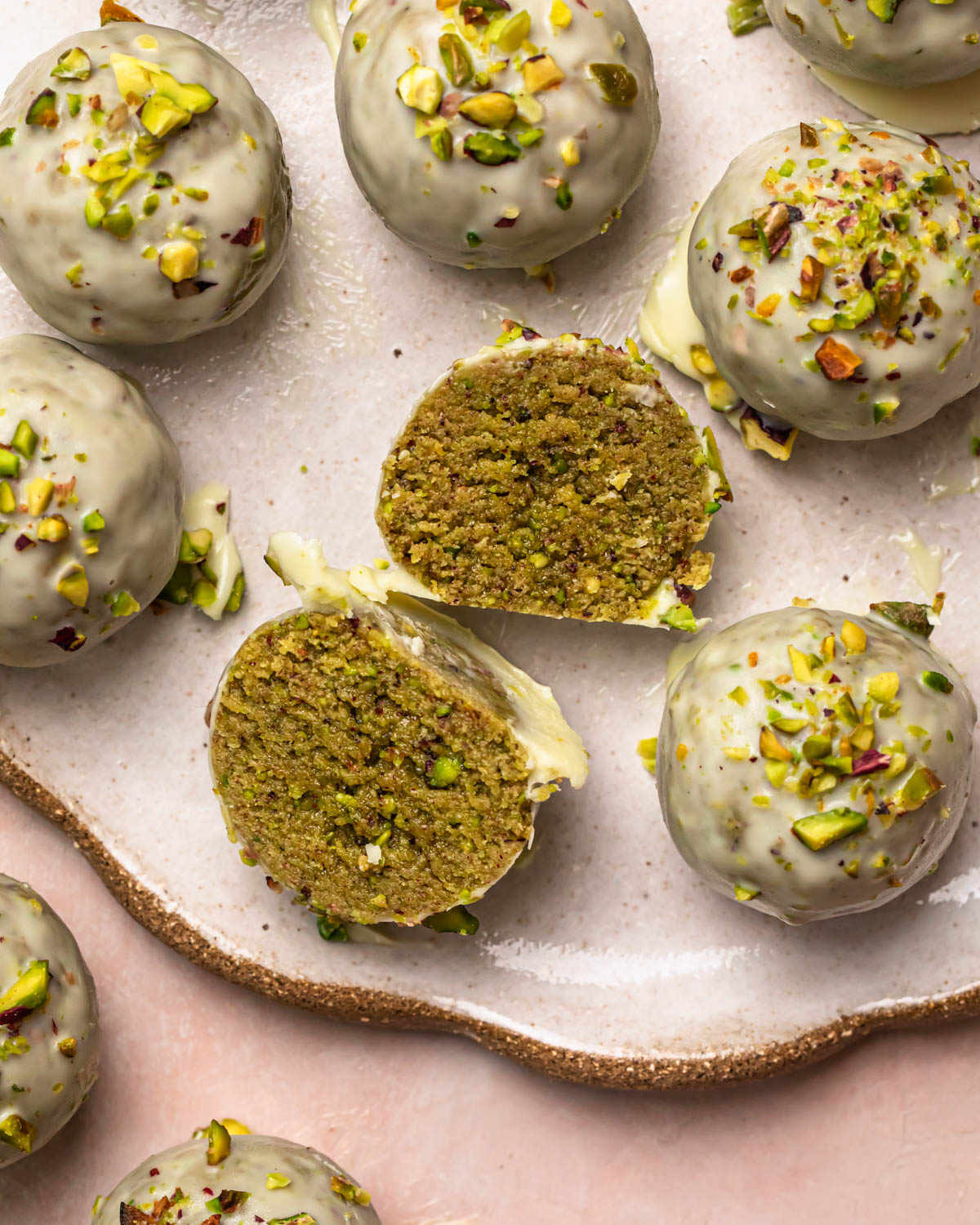 Pistachio protein balls coated in white chocolate and topped with finely chopped pistachios, on a white plate with on cut in half.