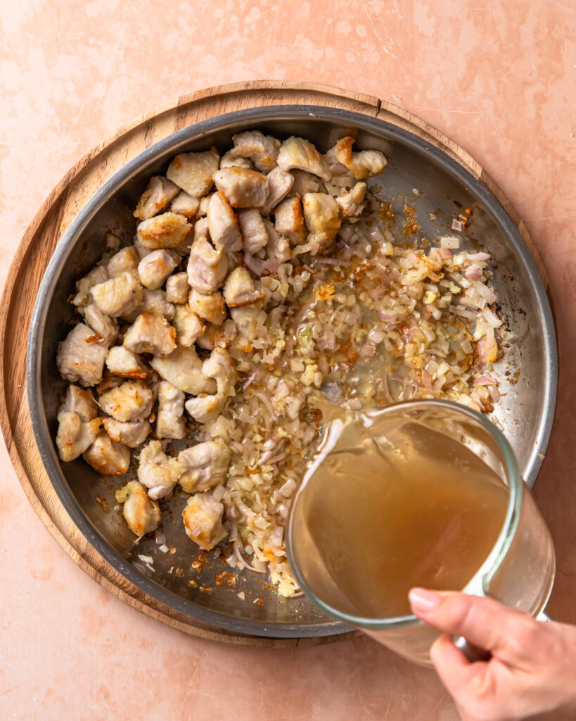 Chicken stock being poured into a pan with chicken and shallots.