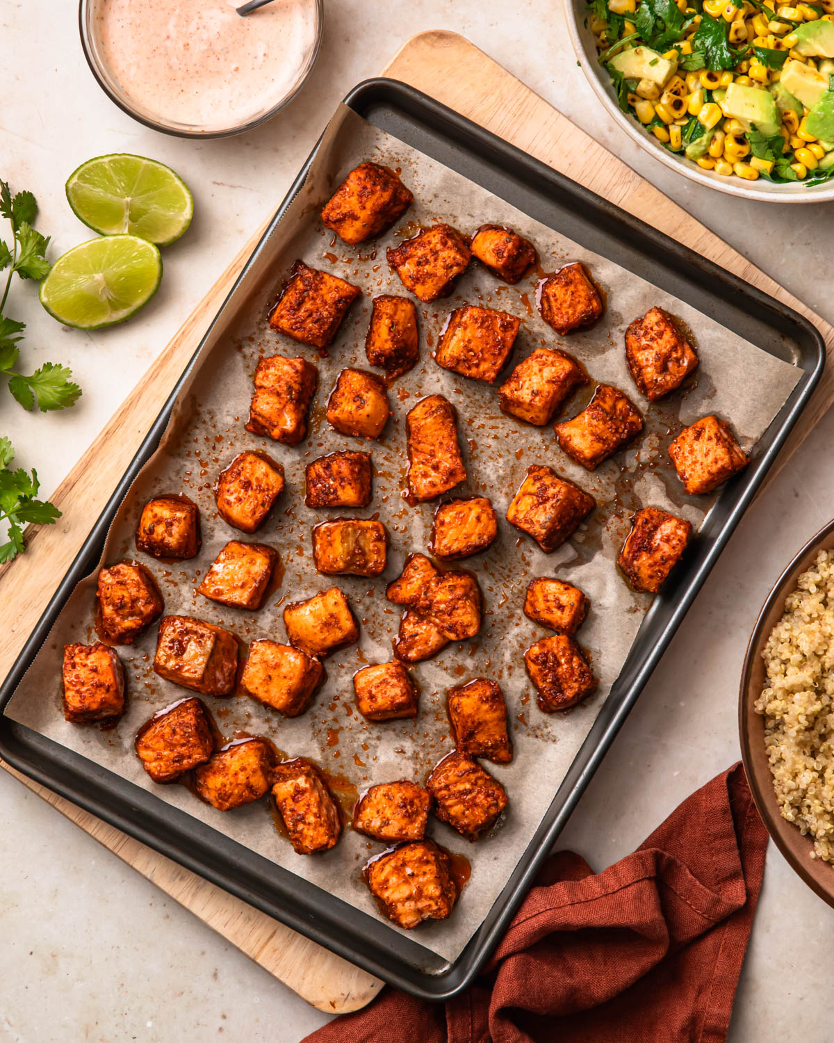 Seasoned and baked salmon bites on a baking tray alongside avocado corn salsa and quinoa. 