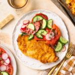Baked parmesan breaded chicken on a plate with a tomato cucumber salad on the side.