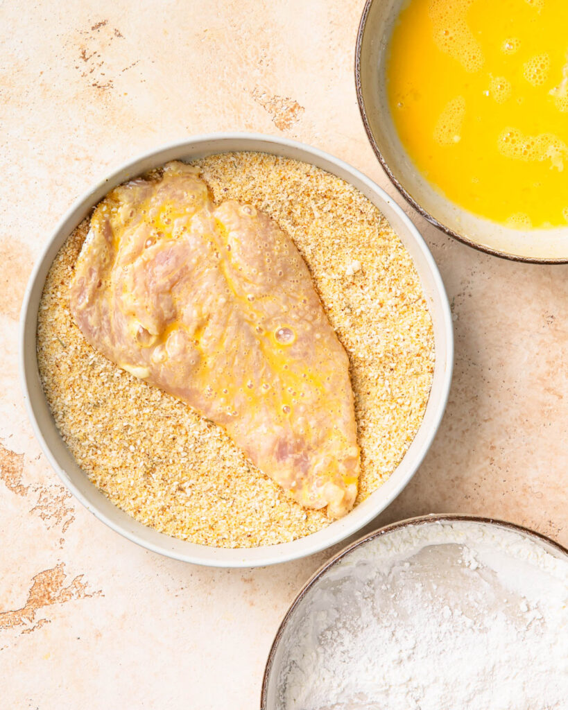 Chicken breast being dipped into a bowl of breadcrumbs.