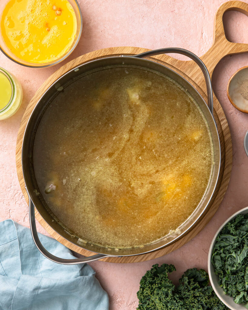 Broth added to a large soup bowl next to a bowl of eggs and a bowl of kale.