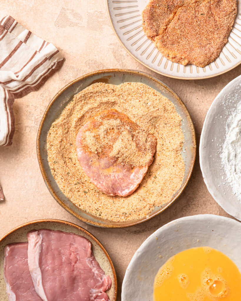 A piece of pork that has been dredged in flour and egg, resting in a bowl of crumb.