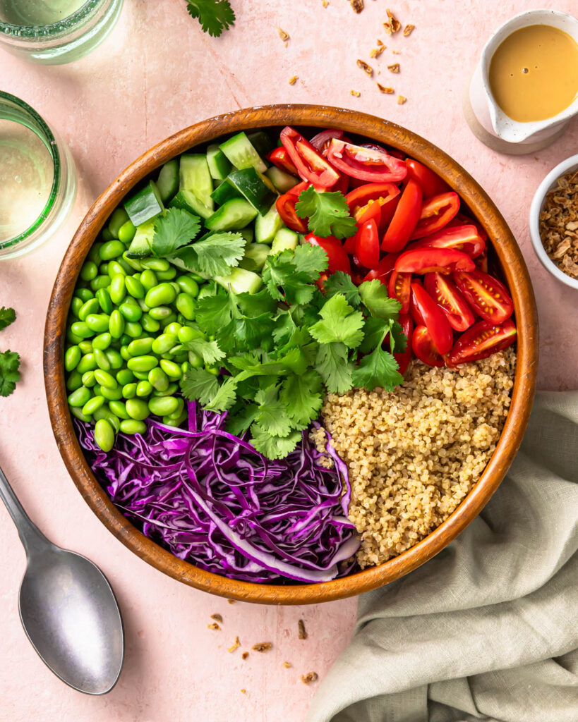 Overhead shot of quinoa, tomatoes, cabbage, edamame, cucumbers, and cilantro in a bowl before mixing.