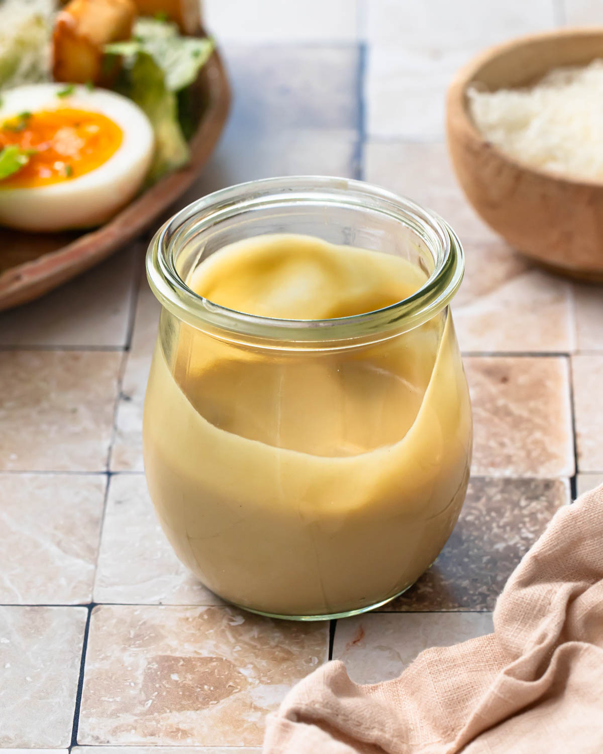 Jar of tahini caesar dressing next to a bowl of parmesan and a salad.