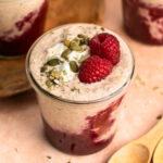 A close up photo of high protein overnight oats in a jar topped with raspberries, yoghurt and pepitas.
