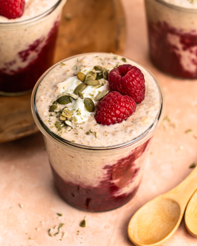 A close up photo of high protein overnight oats in a jar topped with raspberries, yoghurt and pepitas.